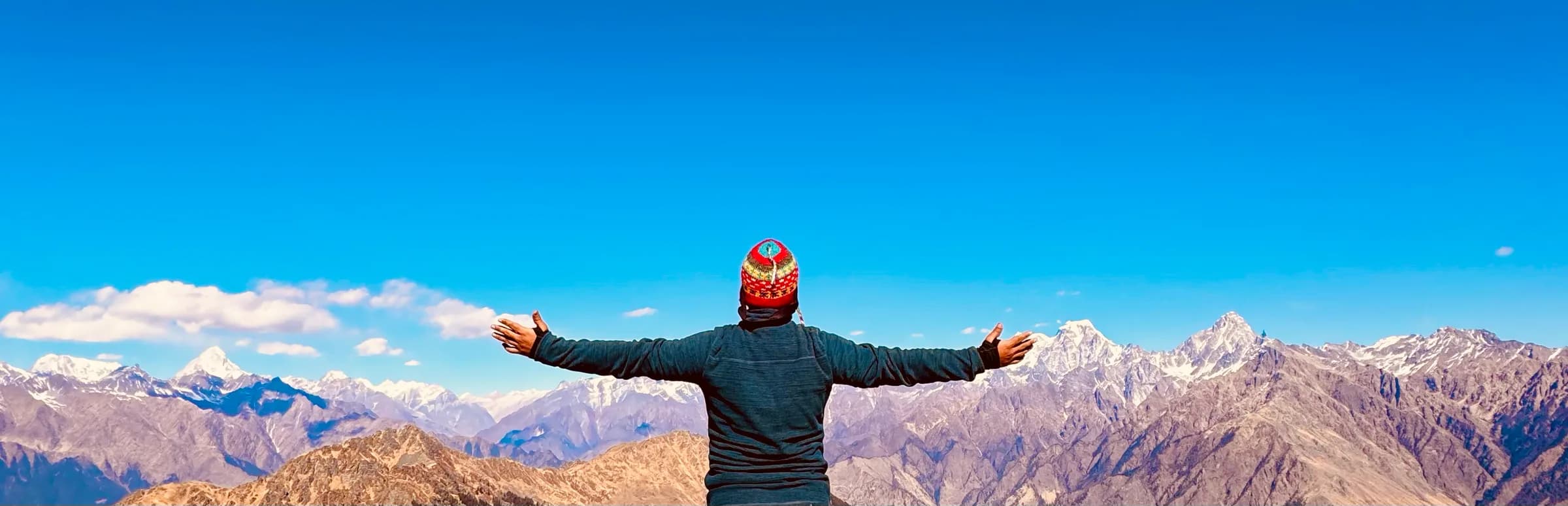Saksham Goyal at a Himalayan summit, arms outstretched toward the snow line